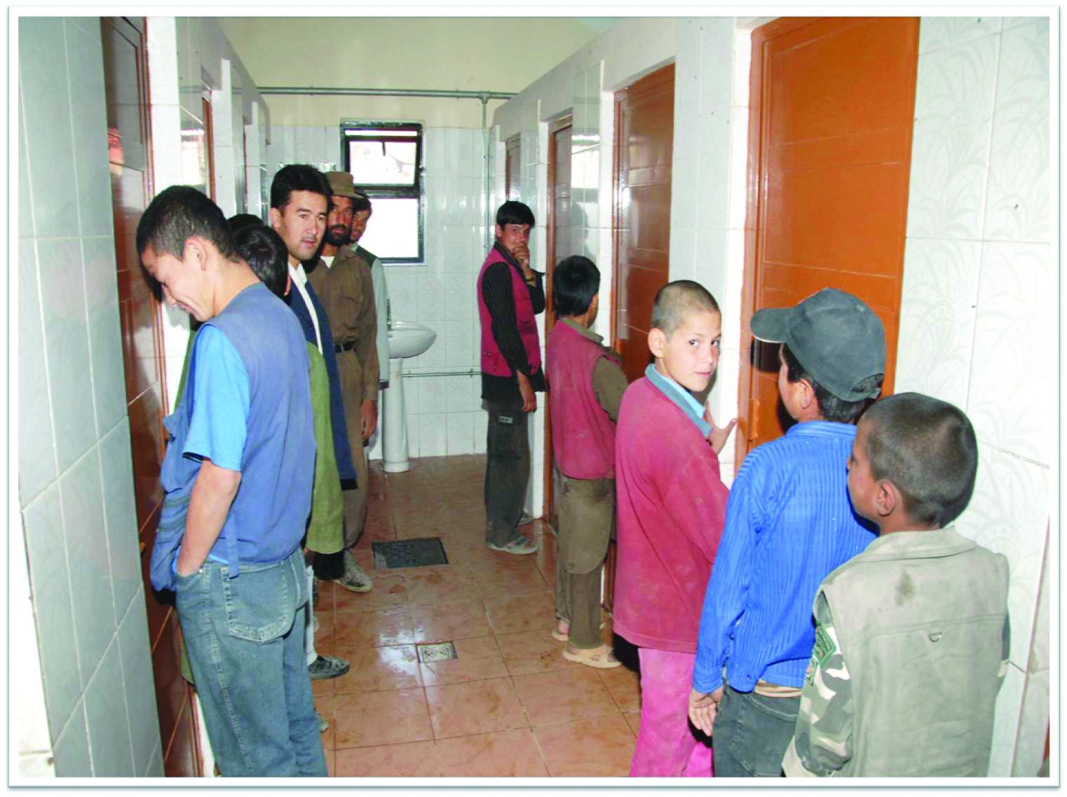 Men and boys waiting inside the tiled washroom of the Kabul complex, with cubicle doors along both walls and a basin at the far end