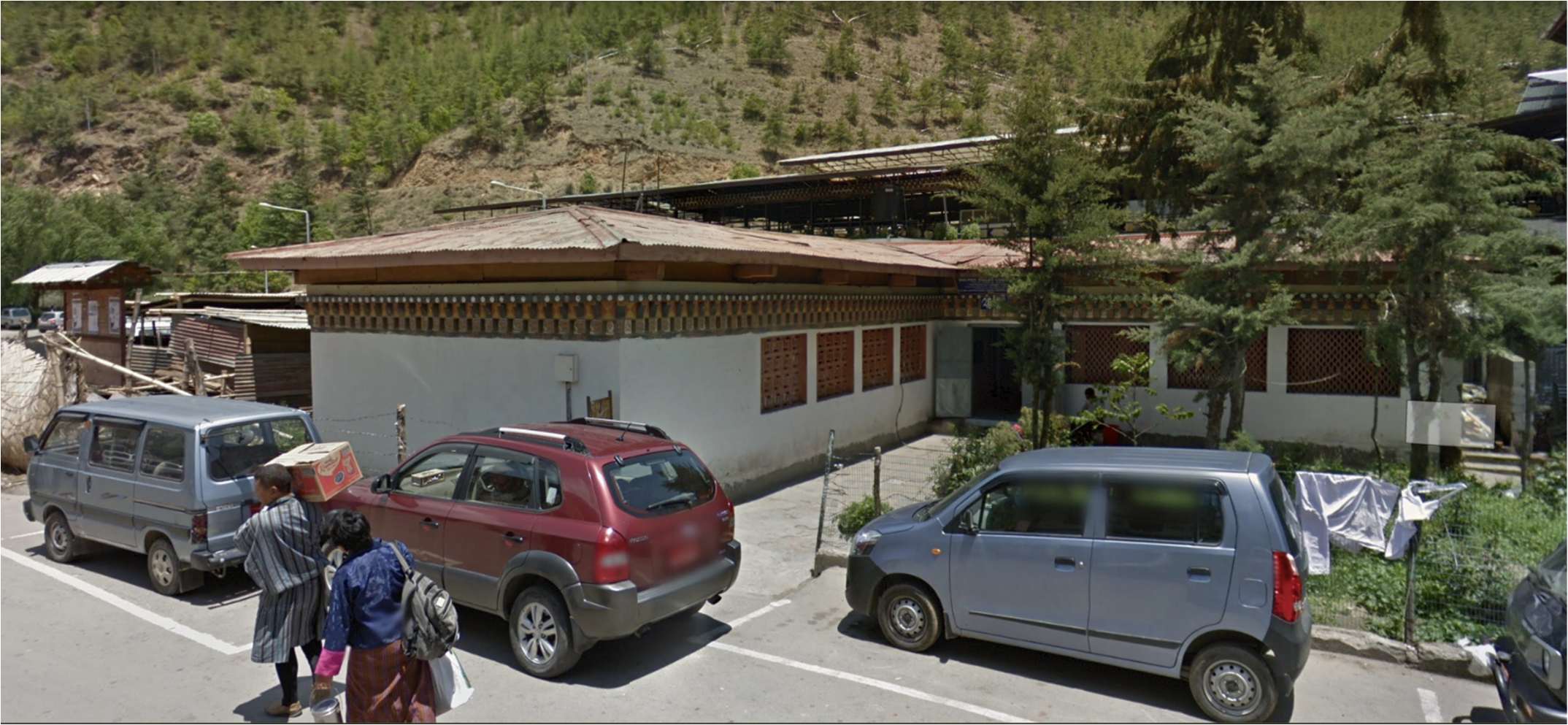 The complex seen from the road, a long single-storey building with a painted timber cornice and a corrugated roof, parked cars in front and forested hills behind
