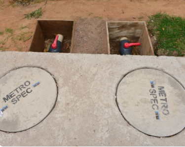 Two circular concrete composter covers stamped Metro Spec, with the open chambers and their valves behind